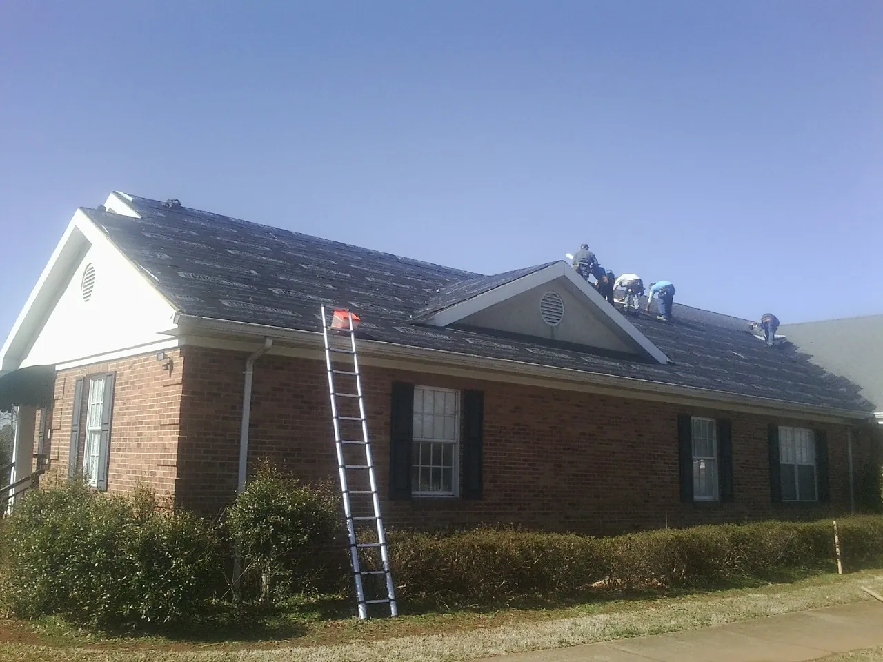 Crew tearing shingles off a brick ranch roof with a ladder set — Emergency Roof Repair work by our crew in Bouton