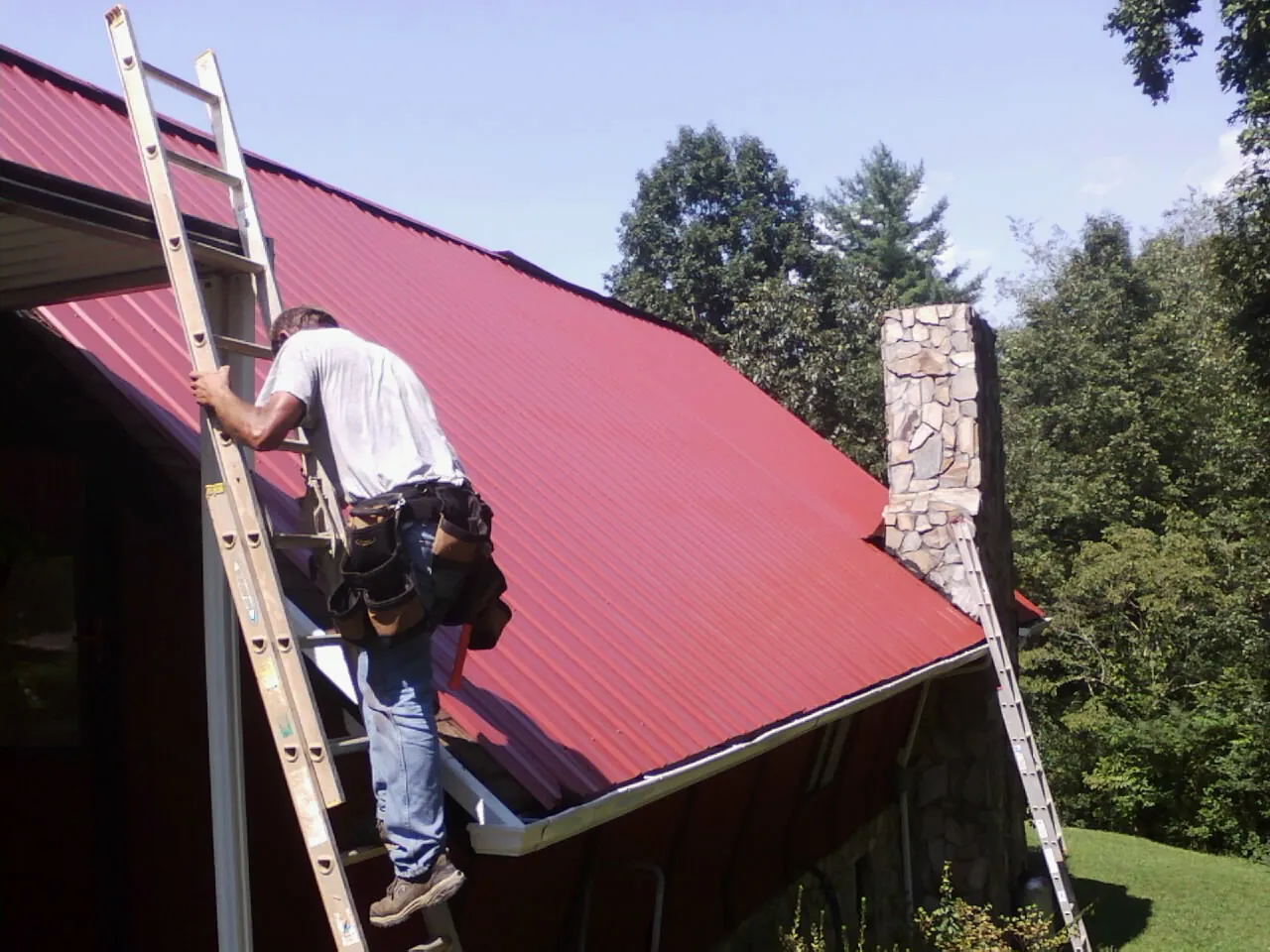 Roofer on a ladder beside a red metal roof and stone chimney — Soffit & Fascia Repair by Roofing Brothers in Bouton